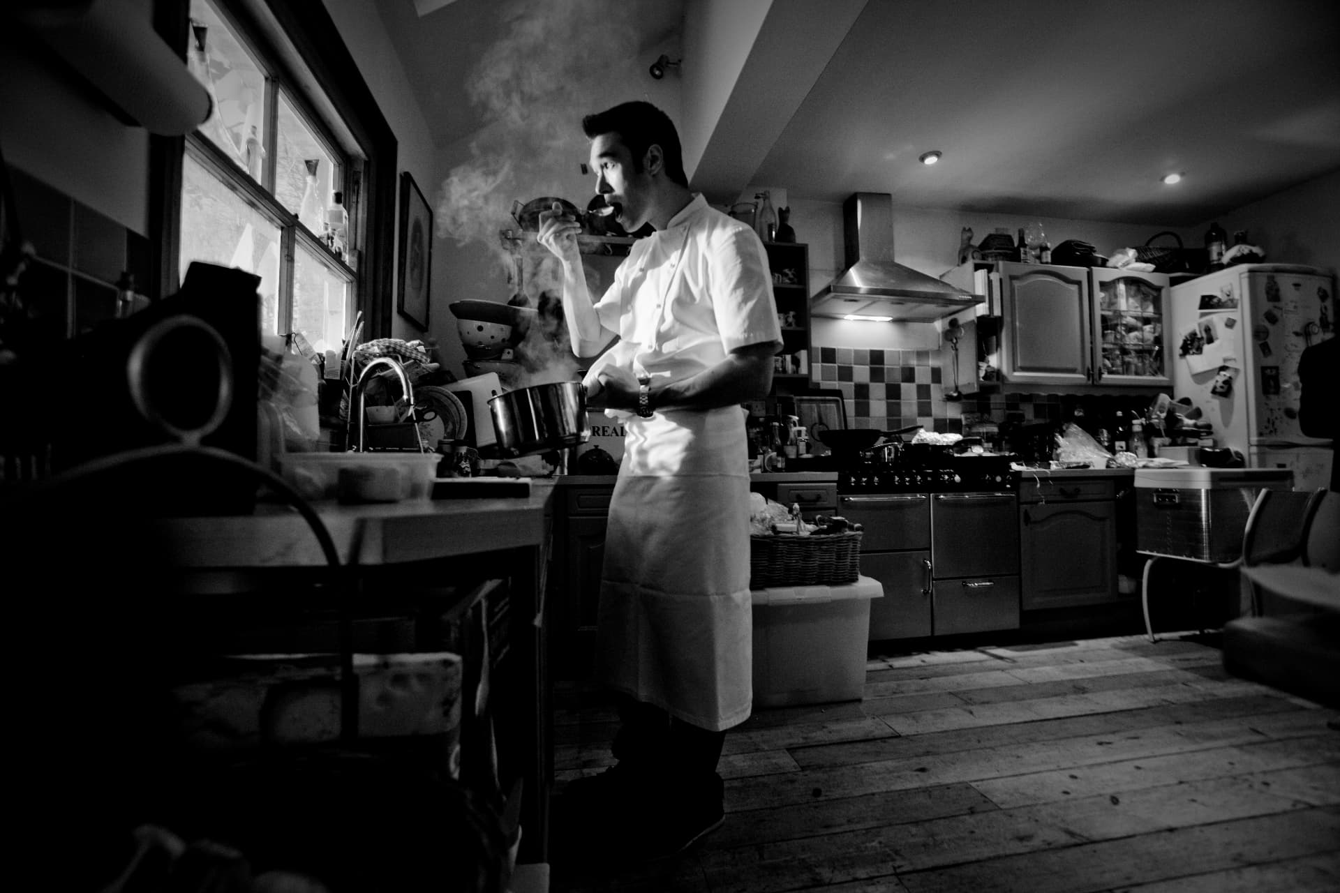 Black-and-white archive photograph of a young Gregory tasting from a pan in a home kitchen.
