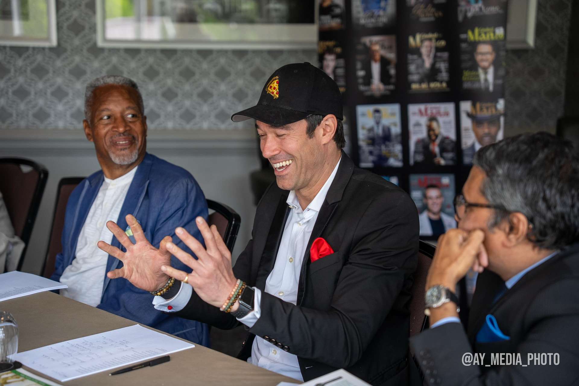 Gregory in a suit jacket speaking animatedly while two men listen across the table.